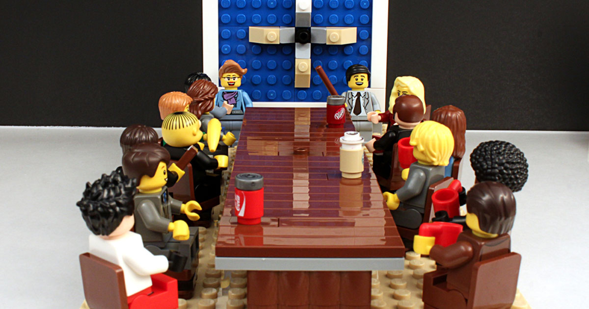 Lego characters sitting at conference table with speaker.