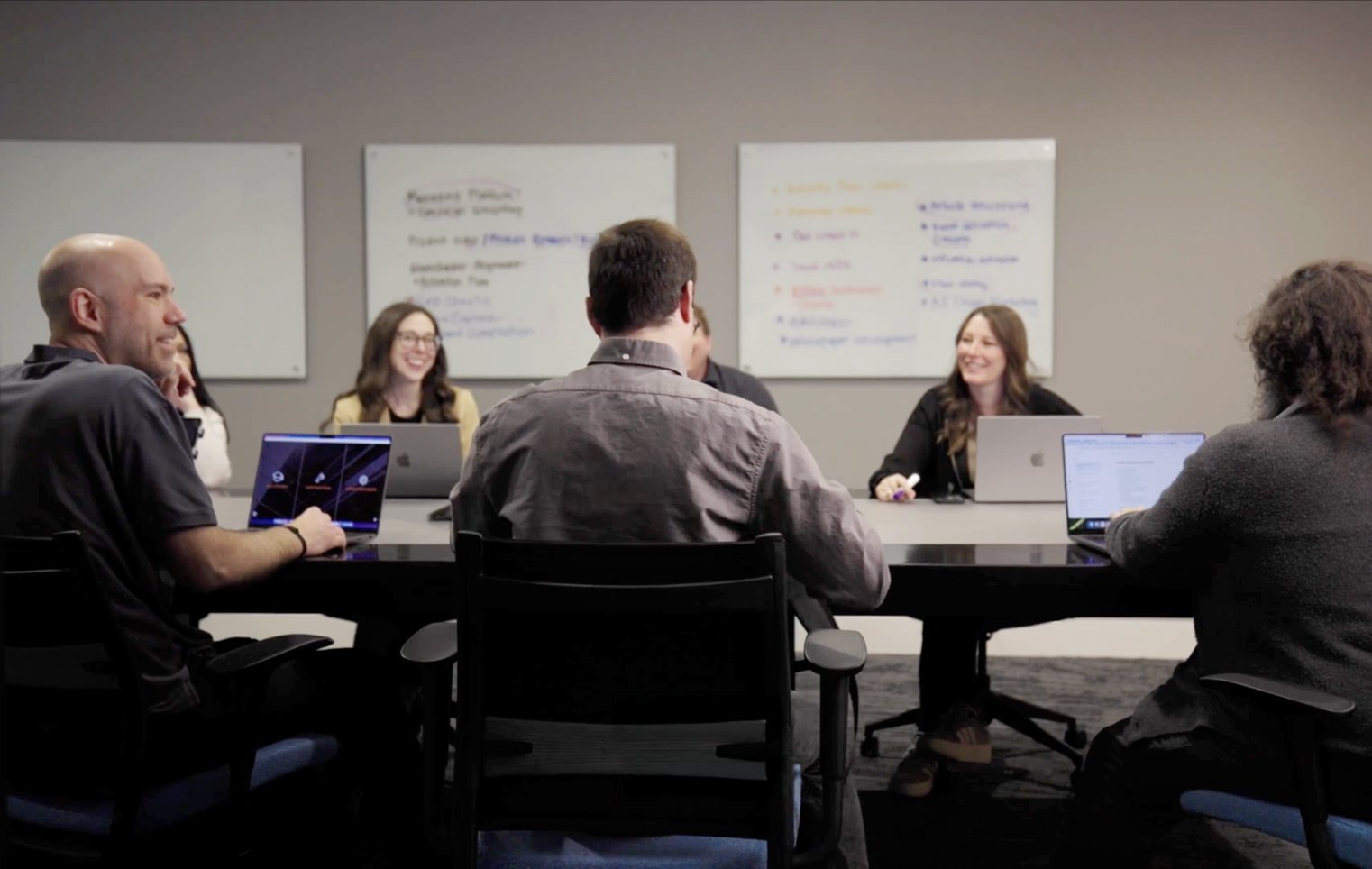 Team members collaborate around a conference table with laptops and whiteboards during a group strategy and planning meeting.