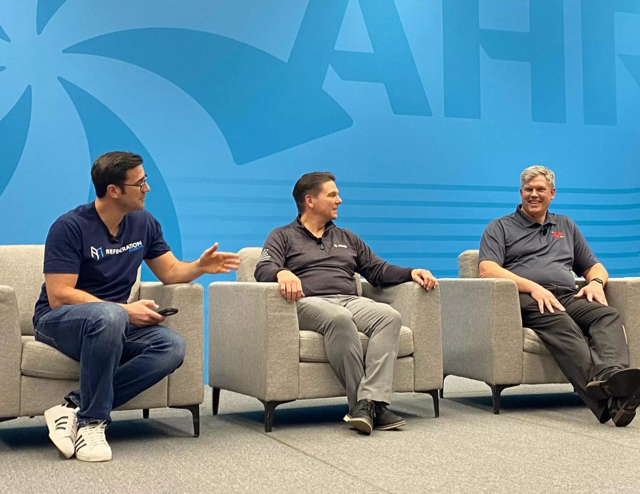 Three HVAC industry professionals seated in armchairs participate in a panel discussion against a blue AHR Expo backdrop.