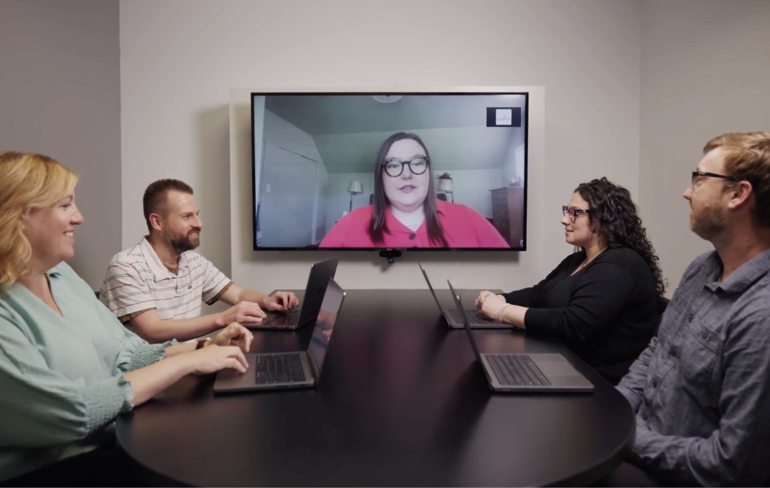 Team members sit around a conference table joining a video meeting with a remote colleague on a large screen, showing hybrid collaboration.
