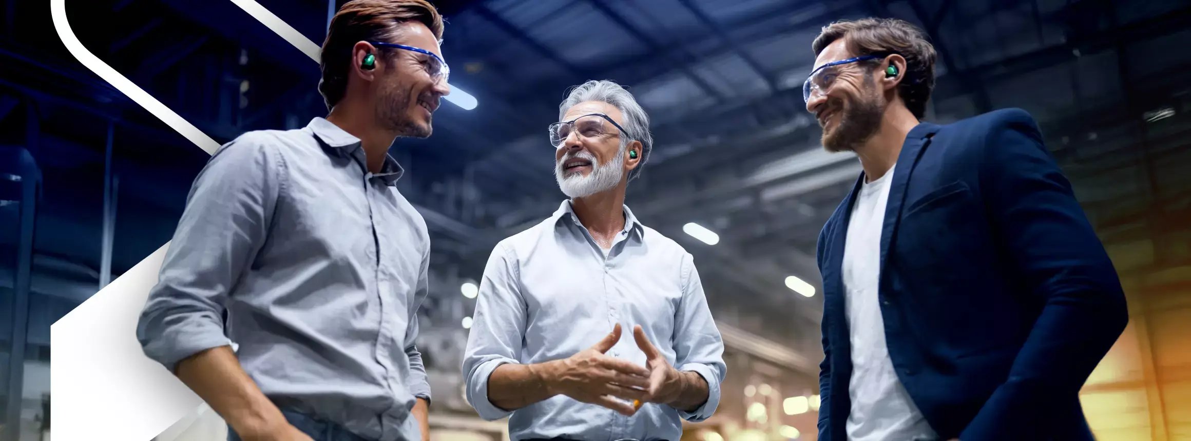 Three professionals wearing safety glasses and hearing protection talk together in a brightly lit industrial facility.