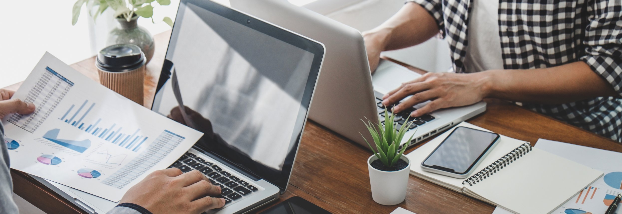Two people work on laptops at a table with printed charts, a smartphone, notebook, coffee cup, and small plant, suggesting business collaboration.