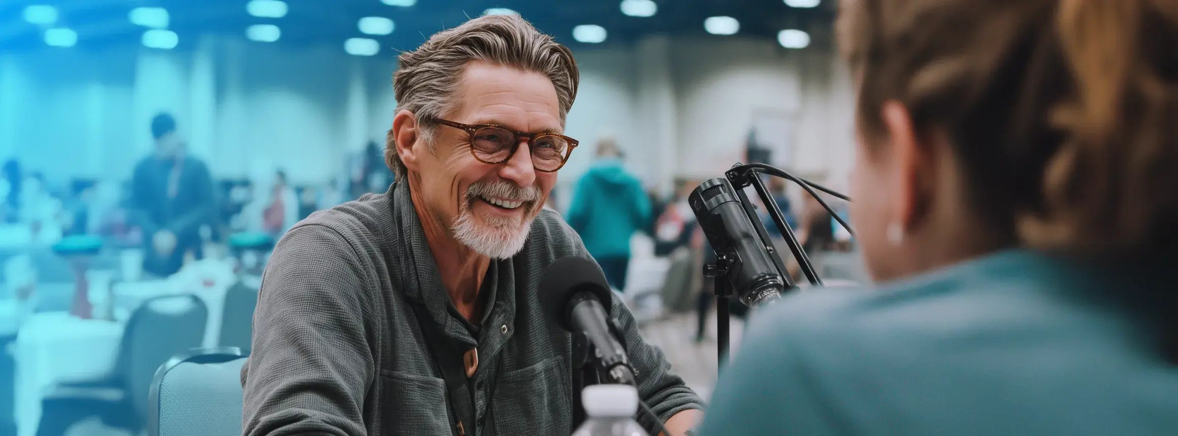 Smiling man with glasses speaks into a microphone during a podcast interview with another person at a busy trade show.
