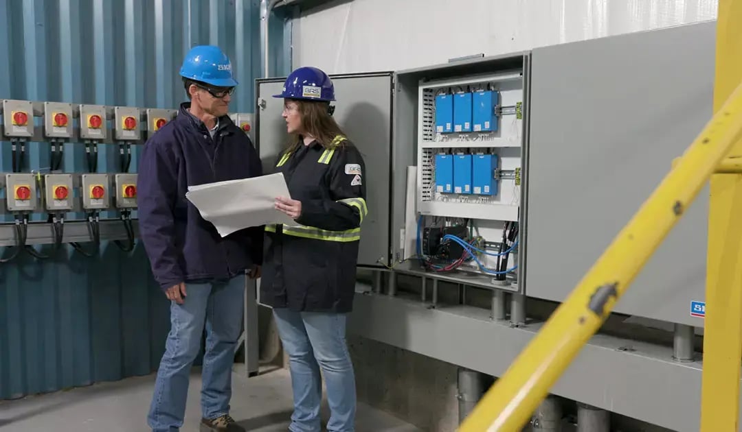 Two workers in hard hats review plans beside an open electrical control cabinet at Big River Steel.