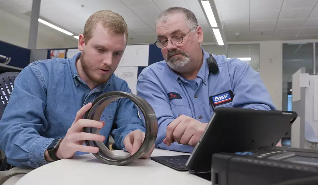 SKF technician and colleague examine a large metal bearing beside a tablet in an office workspace.