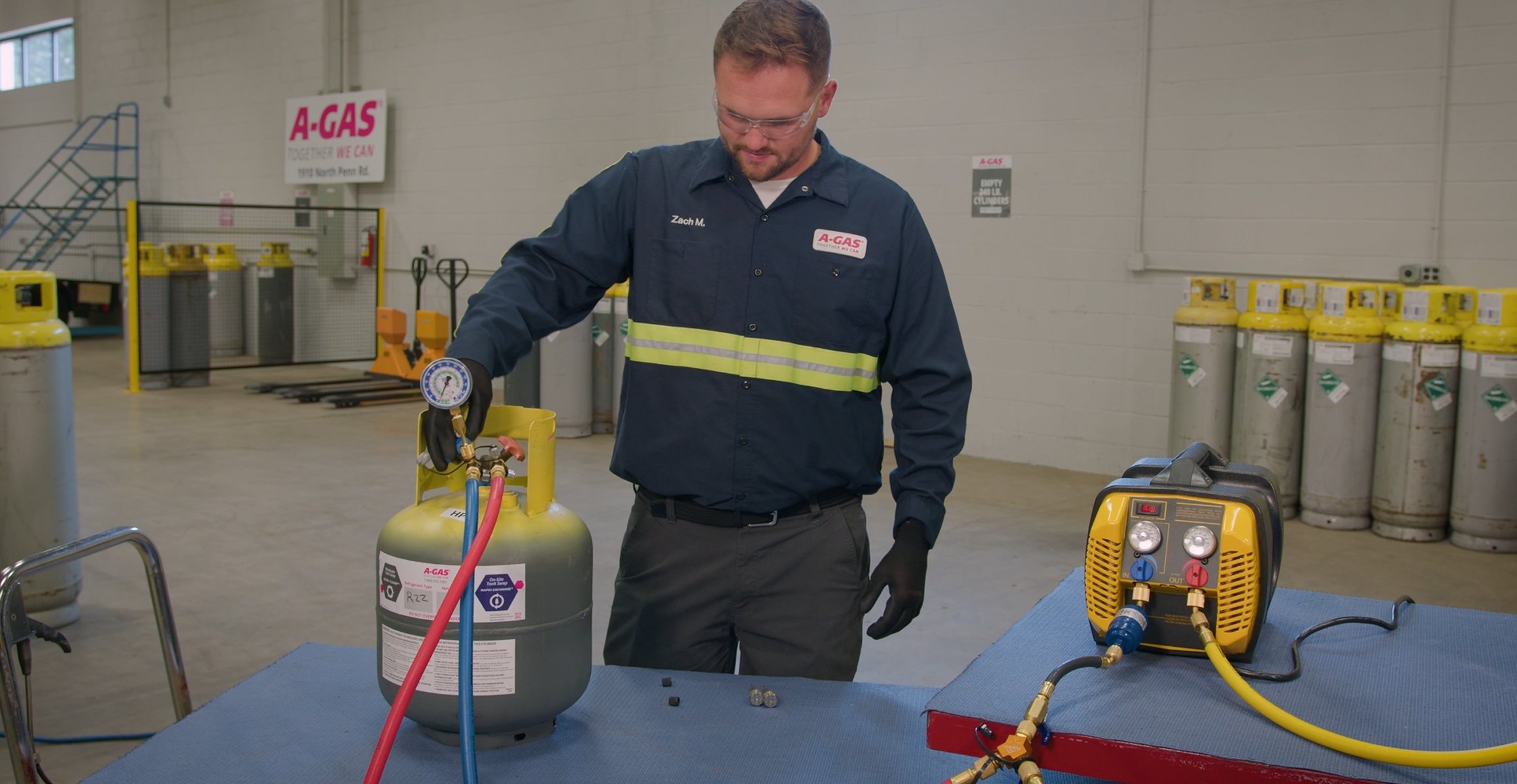 A-Gas technician connects gauges and hoses to a refrigerant recovery cylinder beside recovery equipment in an industrial facility.
