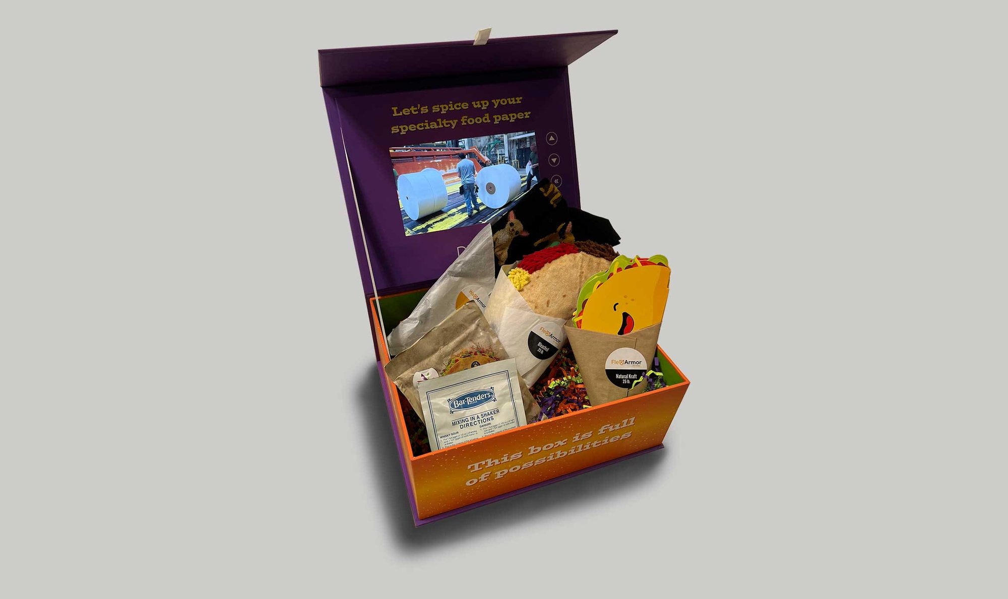 Open Pixelle “Fiesta Fun” box filled with specialty food paper samples, taco-themed items and a screen showing paper production.