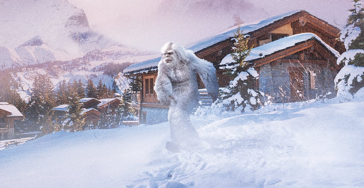 A white yeti walks through deep snow near alpine chalets and mountains, illustrating extreme cold-weather conditions.