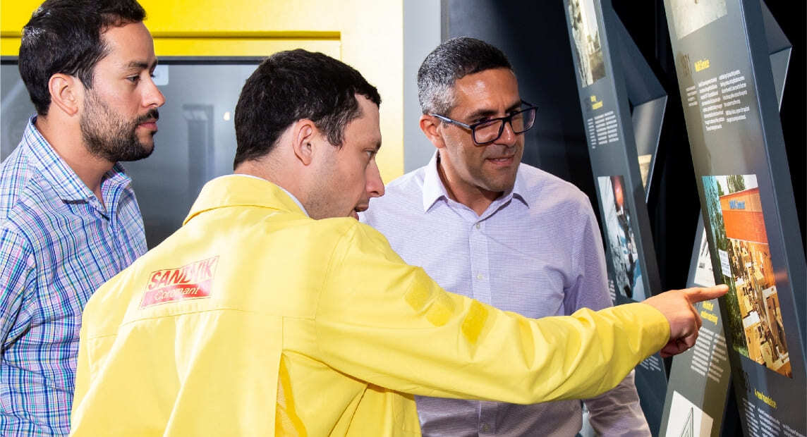 Three men review an informational display, with a Sandvik Coromant employee in a yellow jacket pointing out details.