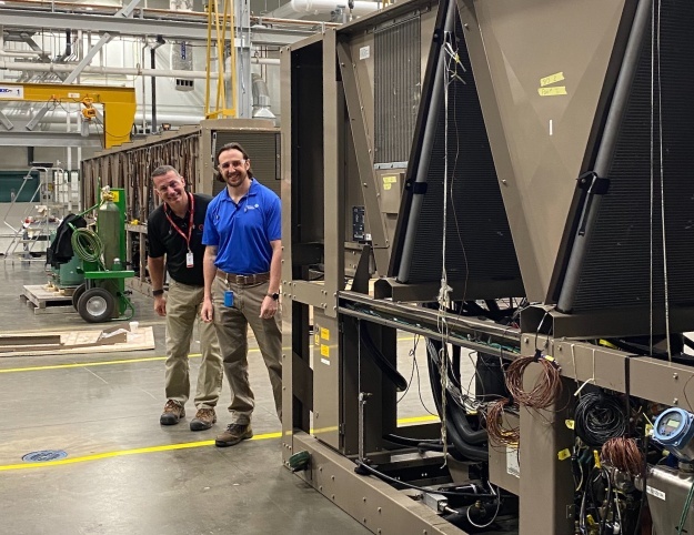 Two HVAC professionals stand beside large commercial HVAC equipment in an industrial testing facility.