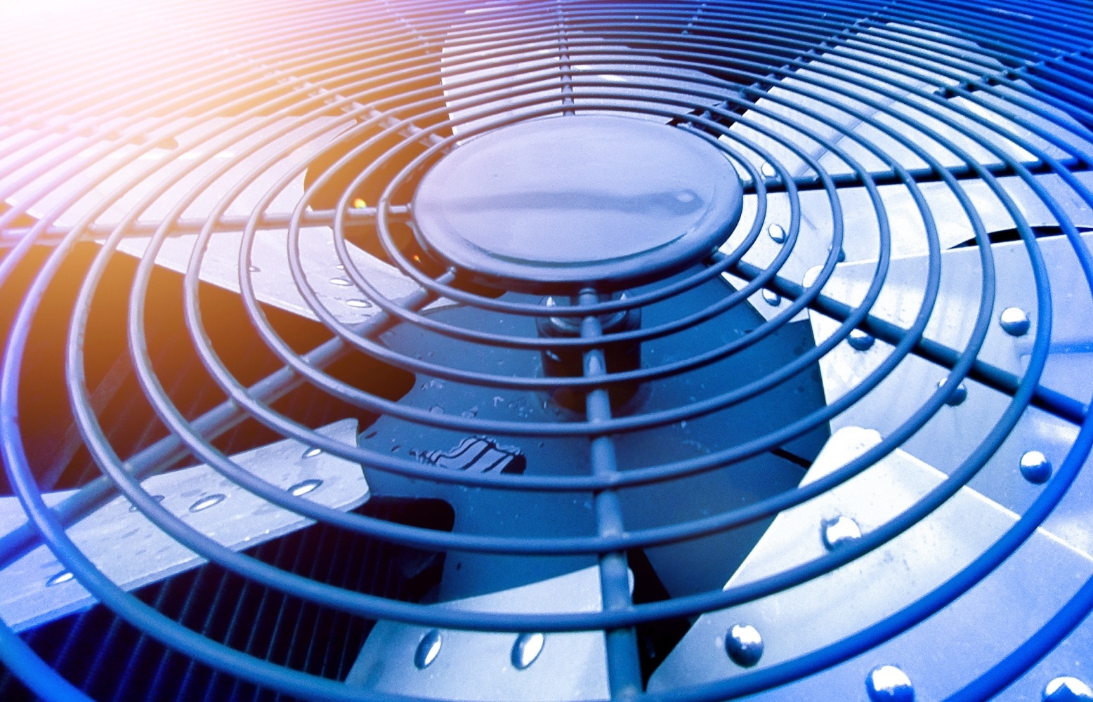 Close-up of an HVAC condenser fan and protective grille with water droplets, illuminated by blue and warm orange light.