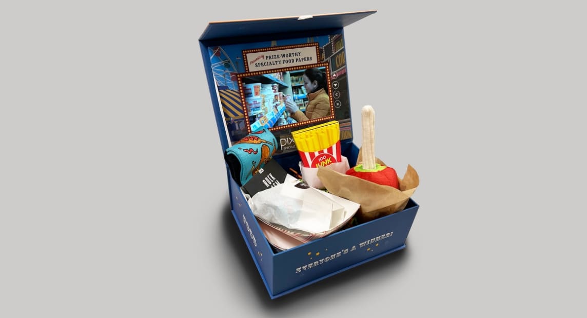 Open Pixelle “Everyone’s a Winner!” box contains carnival-themed props and specialty paper samples beneath a prize-worthy display.