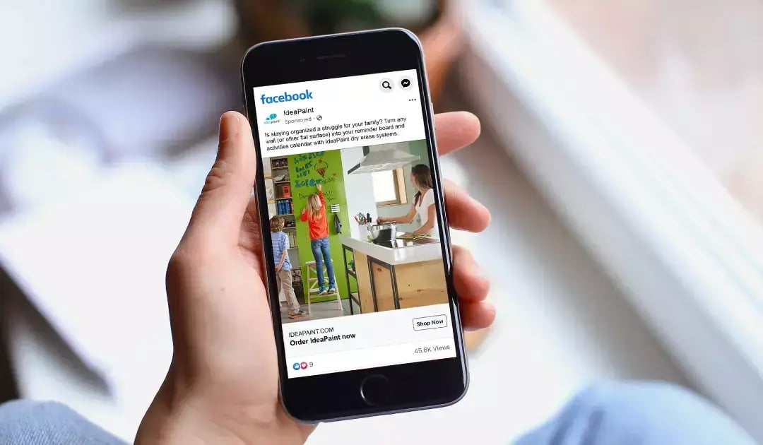 Person holds a smartphone displaying an IdeaPaint Facebook ad showing a family using a dry erase wall.