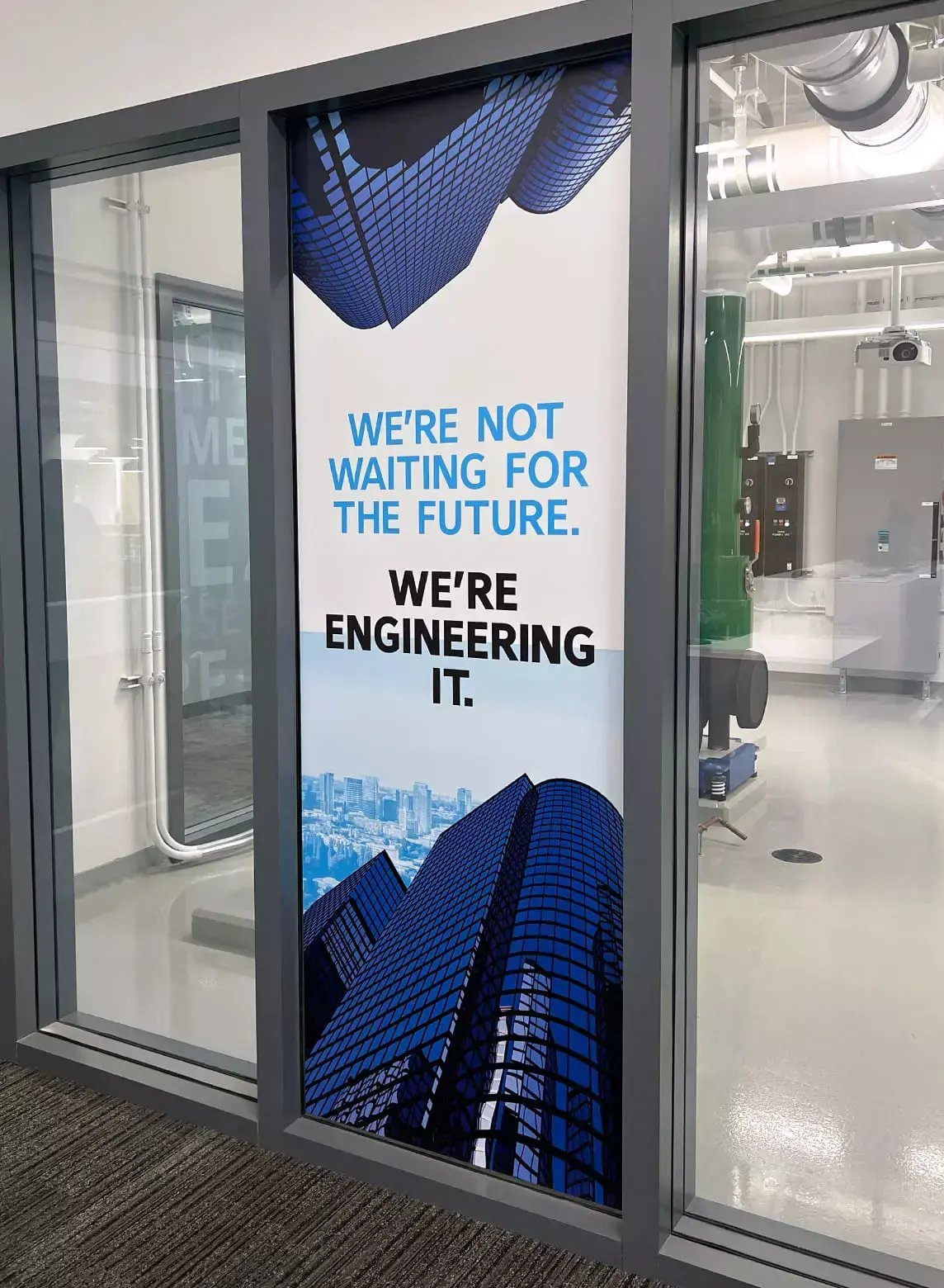 Johnson Controls window graphic at the JADEC facility reads “We’re not waiting for the future. We’re engineering it.” with building imagery.
