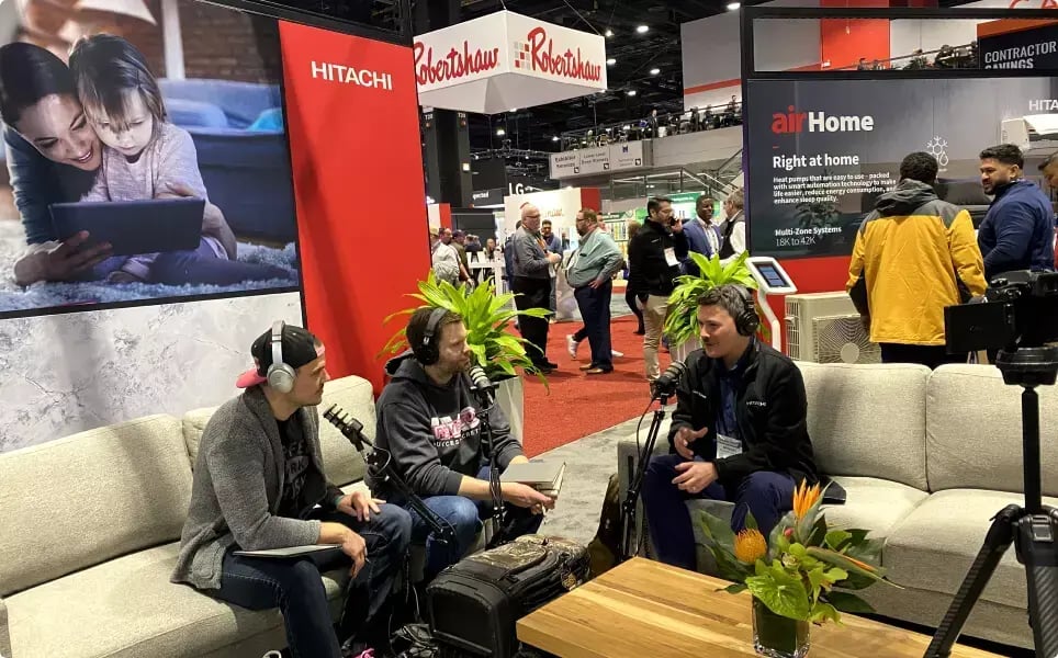 Three participants record an HVAC industry podcast with microphones and headphones at the Hitachi booth during the AHR Expo trade show.