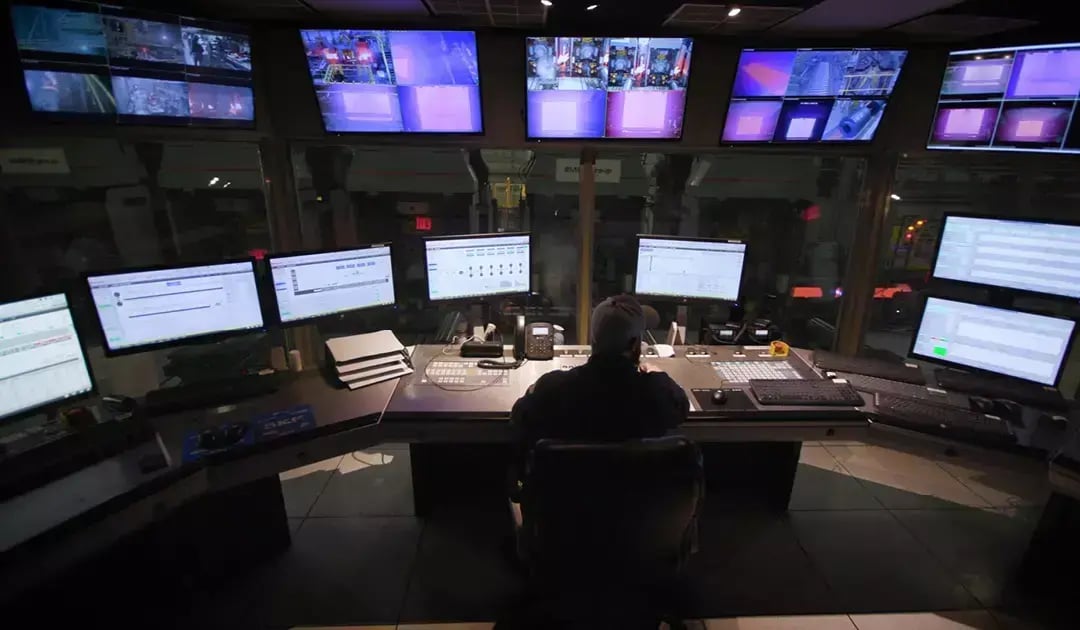 Operator sits at a steel mill control station surrounded by monitors displaying equipment and production data.