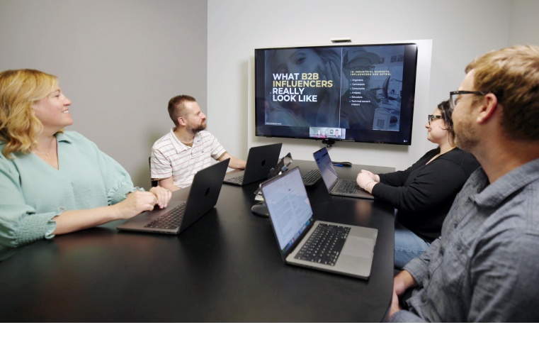 Four colleagues review a presentation titled “What B2B Influencers Really Look Like” on a conference room display during a team meeting.