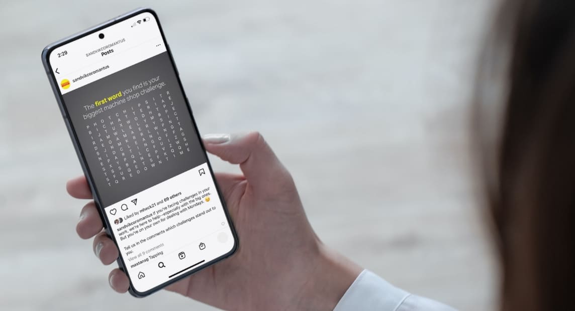 Person holds a phone displaying a Sandvik Coromant word-search post challenging users to find their biggest machine shop challenge.