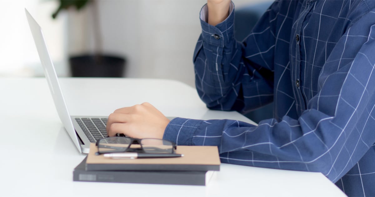 Person in a blue checkered shirt works on a laptop at a desk, with eyeglasses, a notebook and pen resting beside the computer.