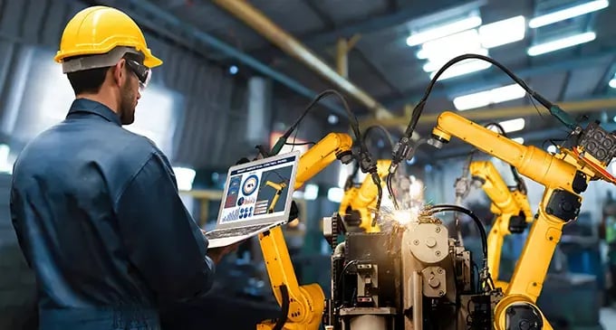 Factory worker monitors robotic welding arms on a laptop as automated machinery welds metal components on a manufacturing line.