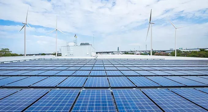 Solar panels cover a large rooftop with several wind turbines in the background, illustrating renewable energy generation.
